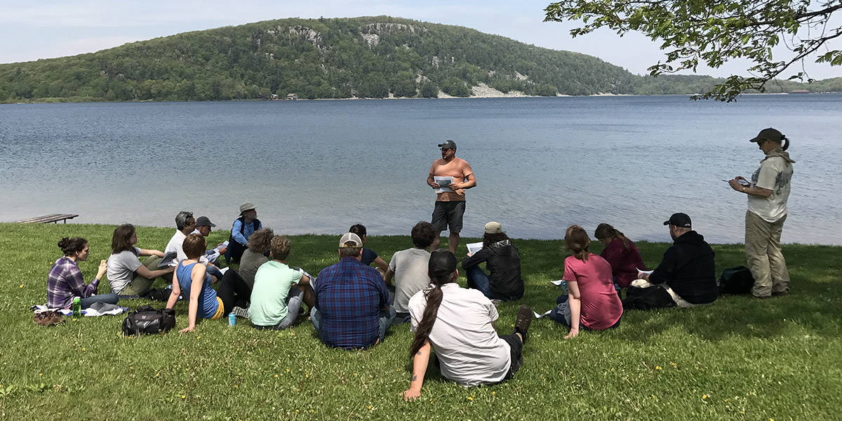 An instructor gives a presentation to a group of students sitting along a lake's edge, with a big hill filling the horizon
