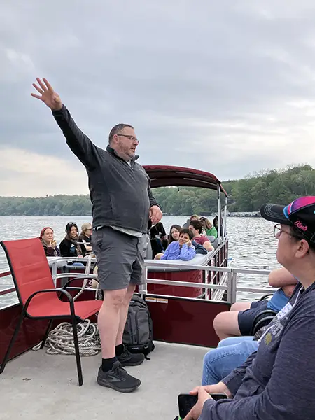 Eric Carson gestures while speaking to a group seated on a pontoon boat on a lake, with trees and cloudy sky in the background.