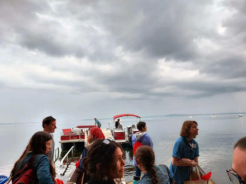 People gathered on a dock under cloudy skies, preparing to board pontoon boats on a calm lake with sailboats in the distance.