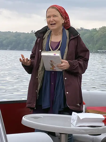 Jane Hirshfield speaks on a pontoon boat while holding a book, with a lake and trees in the background.