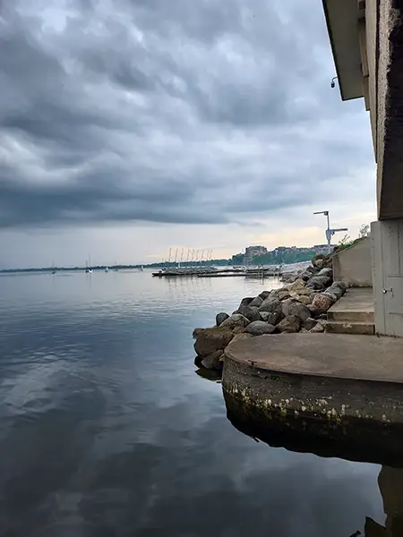 Rocky shoreline and concrete steps along Lake Mendota under dark clouds, with distant sailboats and a pier in view.