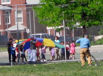 Group of children holding a large rainbow-colored flag while a person rakes grass