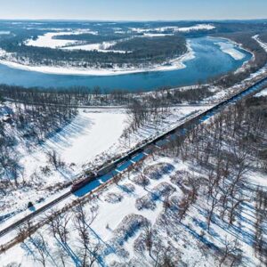 A bird's-eye view of a winter landscape reveals snow-covered terrain, trees, and a train passing near a river.