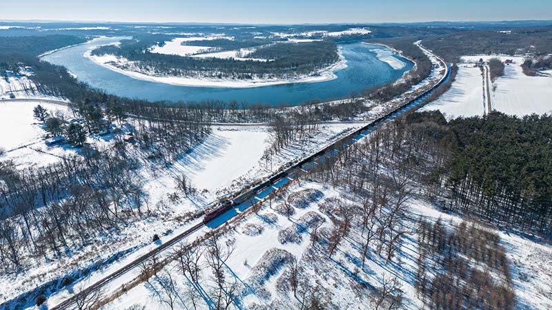 A bird's-eye view of a winter landscape reveals snow-covered terrain, trees, and a train passing near a river.