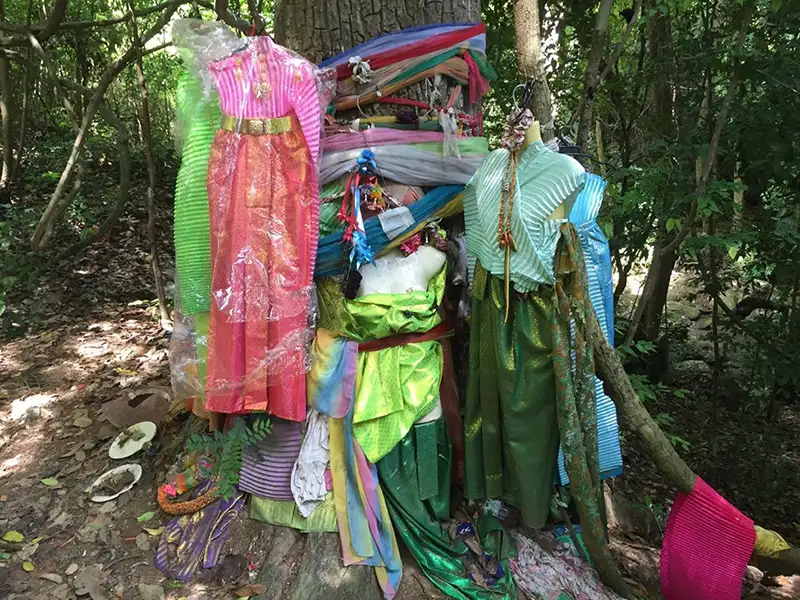 Brightly colored Thai spirit dresses and cloth offerings tied around a tree trunk in a forest shrine.